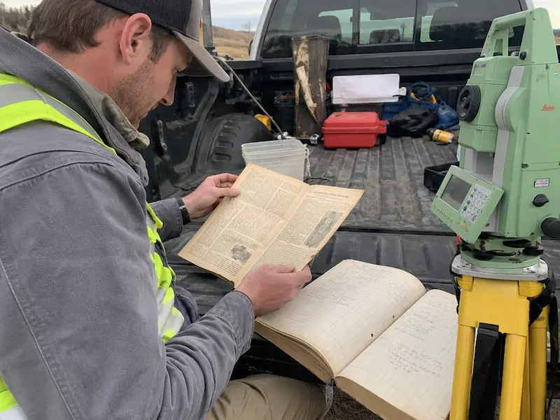 Modern land surveyor reading a historic surveying article with field book and total station nearby
